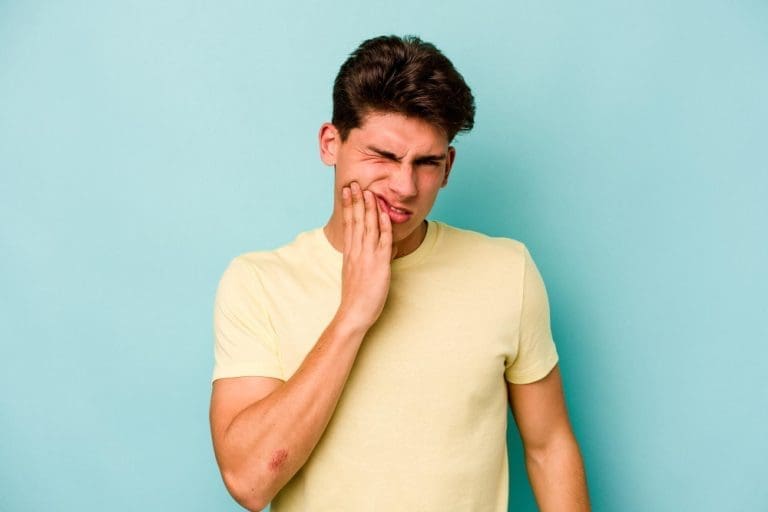 A young man in a yellow t-shirt stands against a blue background, wincing and holding his cheek—a reminder that urgent dental issues can arise unexpectedly. Stellar Orthodontics in San Marcos, CA offers emergency orthodontic care for sudden toothaches or facial discomfort.