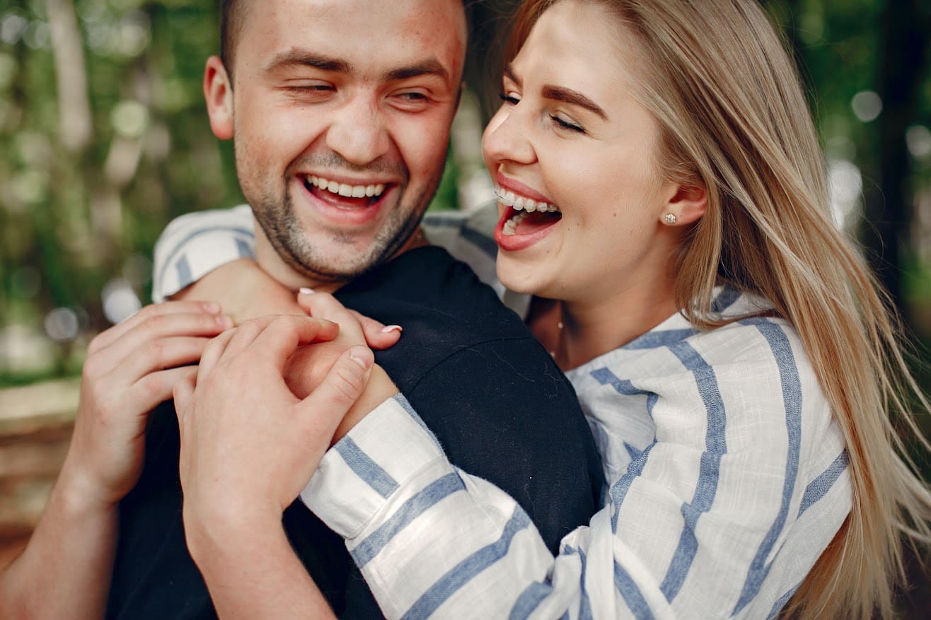 A man and woman smile and laugh together outdoors, embracing with blurred trees in the background—capturing the joy you'll experience with a complimentary consultation at Stellar Orthodontics in San Marcos, CA.
