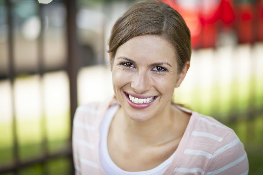 A woman with light brown hair and a light-colored striped top smiles at the camera, photographed with digital imaging by Stellar Orthodontics in San Marcos CA, against a softly blurred outdoor background.