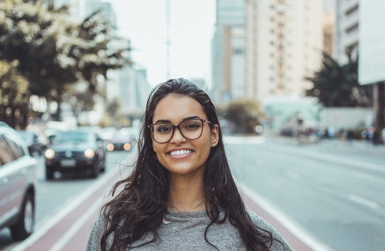 A woman with long dark hair and glasses smiles, showing off her Smile Touch Up from Stellar Orthodontics in San Marcos CA, while standing on a city street lined with cars and tall buildings.