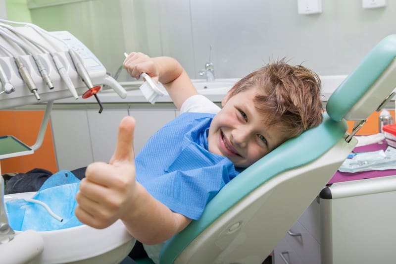 At Stellar Orthodontics in San Marcos CA, a boy sits in the dentist chair wearing a blue bib, smiling and giving a thumbs up to the camera. Nearby dental equipment highlights their expertise in children's orthodontics.