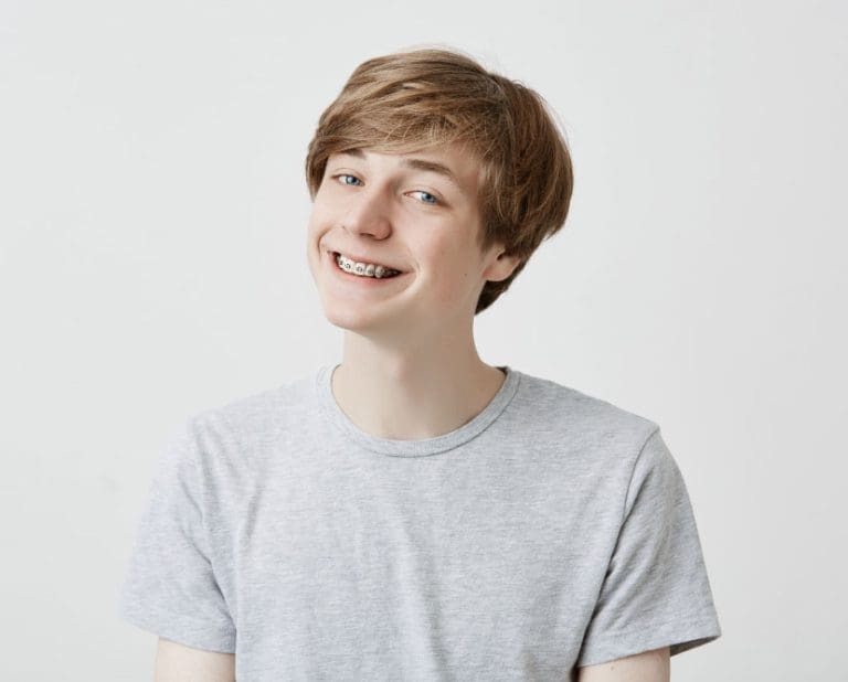 A smiling teen with light brown hair, braces, and a grey t-shirt poses against a white background—perfect for showcasing Treatment For Teens at Stellar Orthodontics in San Marcos, CA.