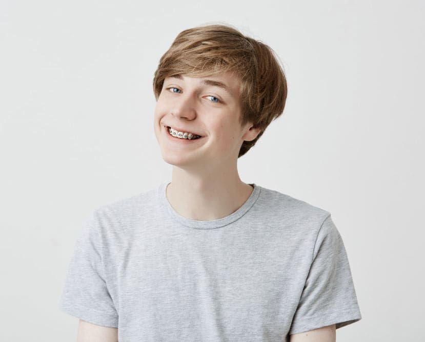 A smiling teen with light brown hair, braces, and a grey t-shirt poses against a white background—perfect for showcasing Treatment For Teens at Stellar Orthodontics in San Marcos, CA.