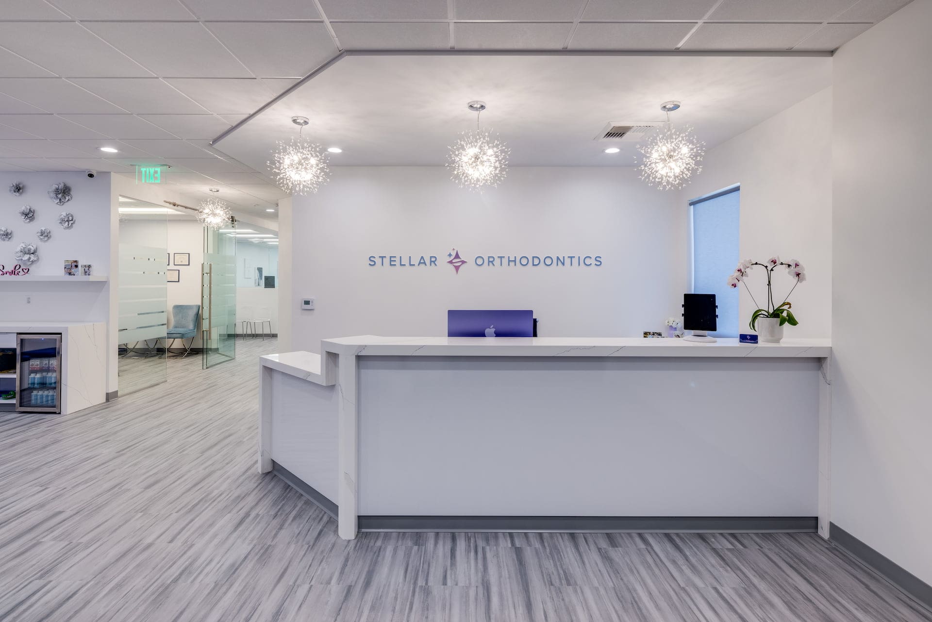 The reception area at Stellar Orthodontics in San Marcos, CA features a white desk, modern lighting, an orchid plant, and a wall sign behind the counter.