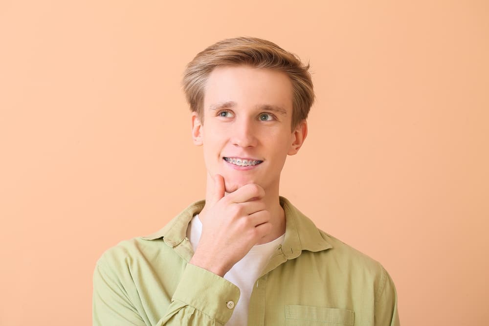 At Stellar Orthodontics in San Marcos CA, a young man with light brown hair and clear braces wears a green shirt and stands against a peach background, looking thoughtfully to the side.