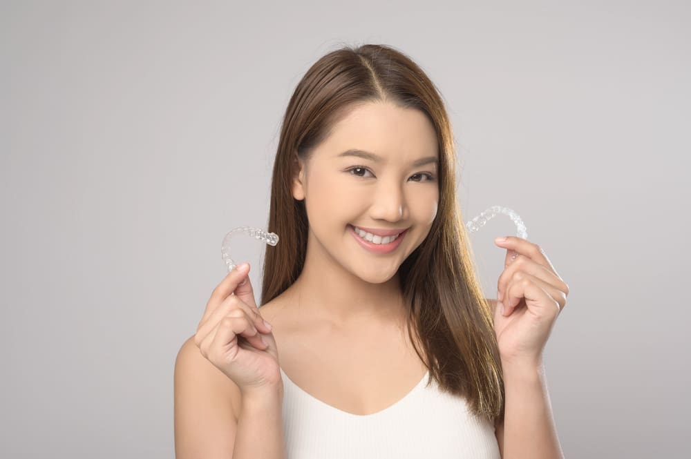 A woman with long brown hair holds Invisalign clear aligners and smiles in San Marcos CA, representing Stellar Orthodontics against a plain background.