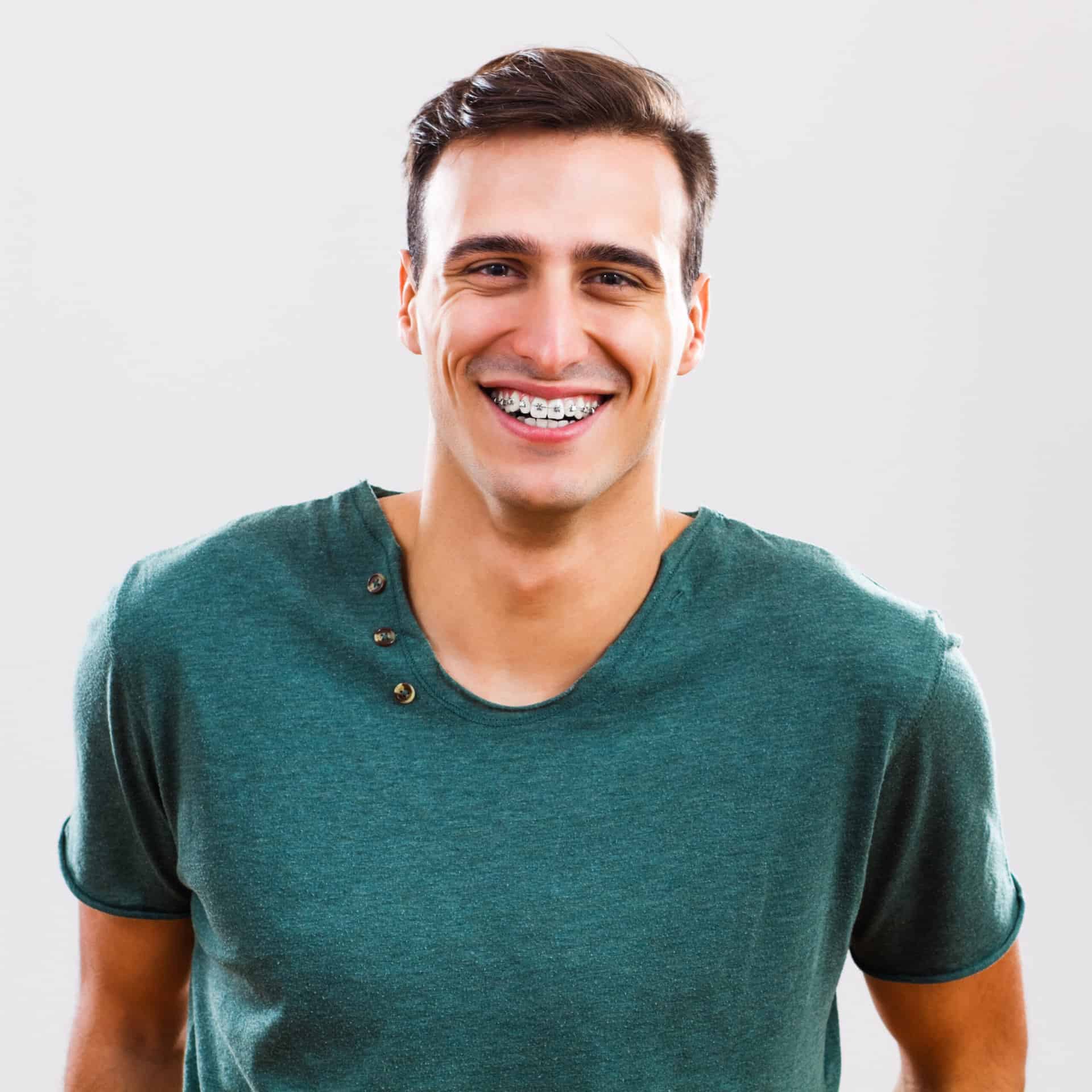 Smiling at the camera in a green T-shirt, a young man with short brown hair confidently displays his adult braces from Stellar Orthodontics in San Marcos, CA against a plain light background.