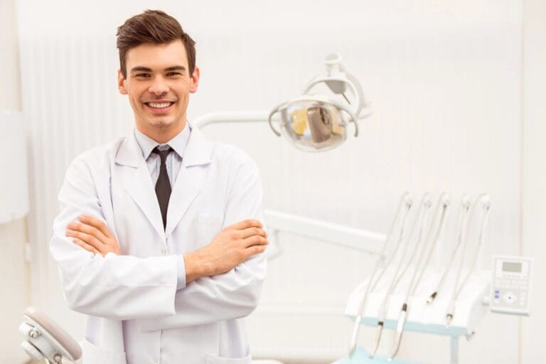 At Stellar Orthodontics in San Marcos, CA, a male dentist in a white coat stands confidently with arms crossed, surrounded by dental equipment and a treatment chair after explaining about How to Fix Overbite in San Marcos, CA.