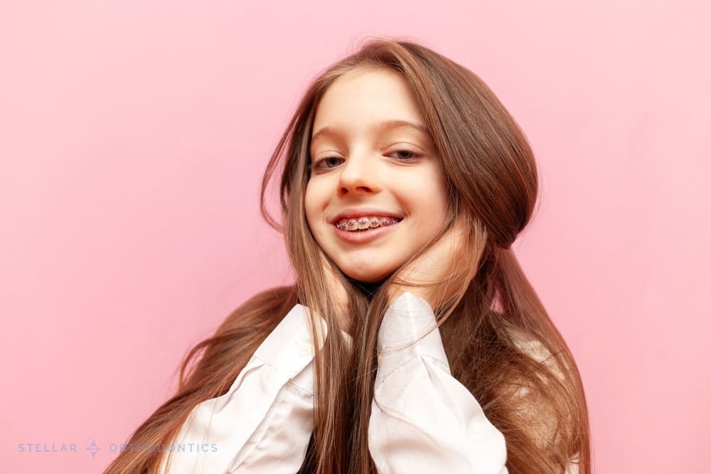 At Stellar Orthodontics, a young girl with long brown hair and braces for overbite in San Marcos, CA smiles brightly, hands on her cheeks, against a pink background.