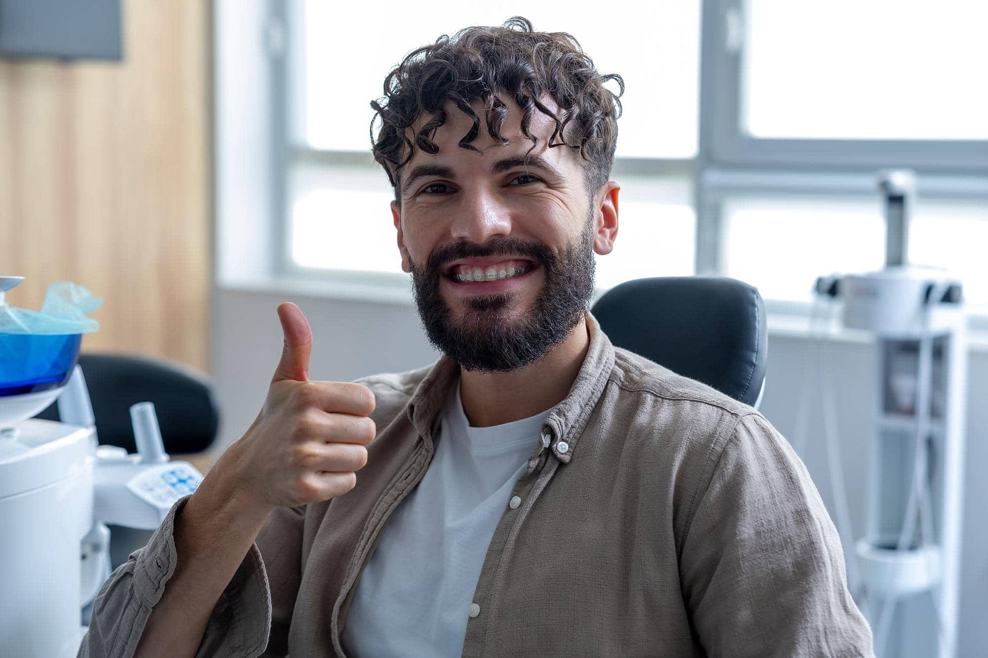 Smiling man with curly hair and clear braces a beard gives a thumbs up in an office at Stellar Orthodontics in San Marcos, CA.