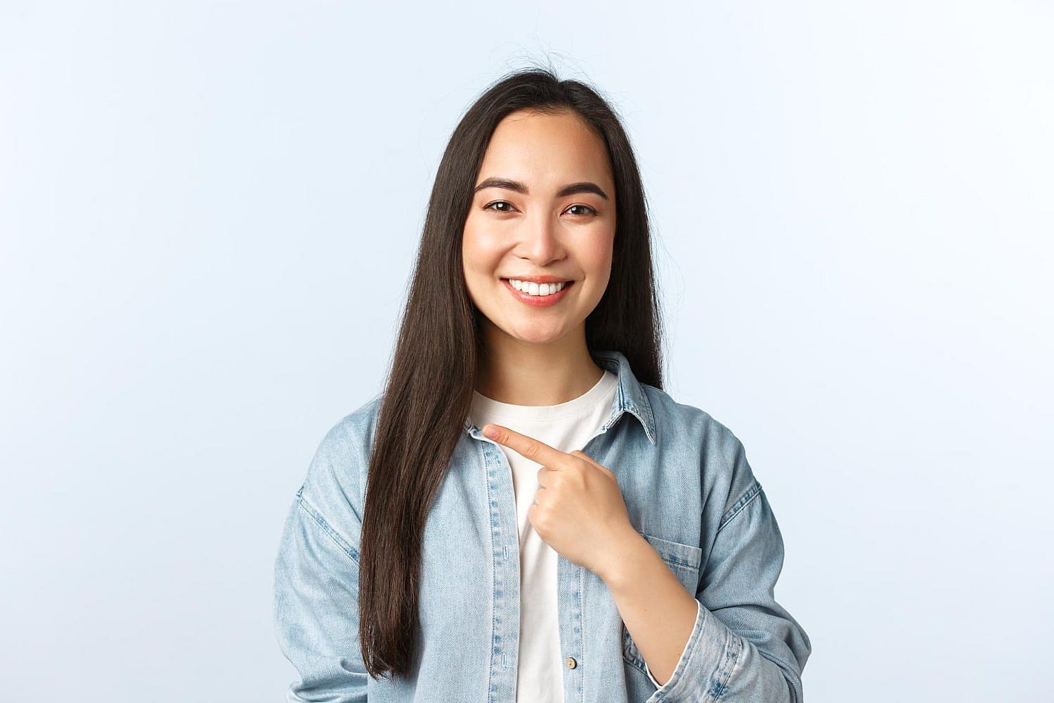 Smiling young woman with long dark hair in a denim shirt points at herself, highlighting Invisalign Treatment at Stellar Orthodontics in San Marcos, CA.