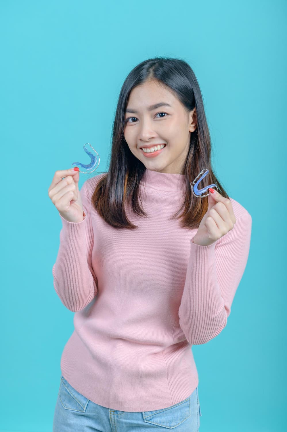 Smiling, a woman in a pink sweater shows two clear dental retainers, orthodontic appliances from Stellar Orthodontics in San Marcos, CA.