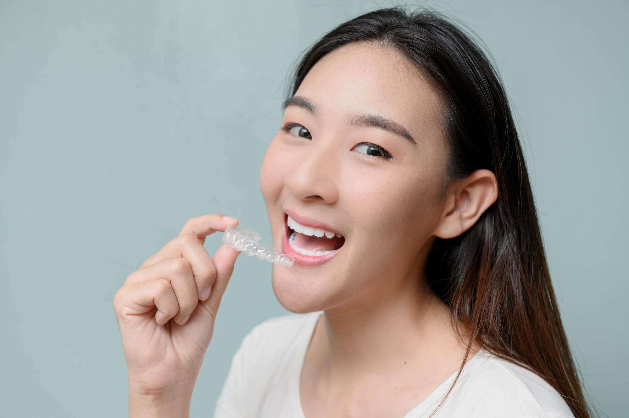 A woman smiles, holding a clear dental aligner Invisalign near her mouth at Stellar Orthodontics in San Marcos, CA.