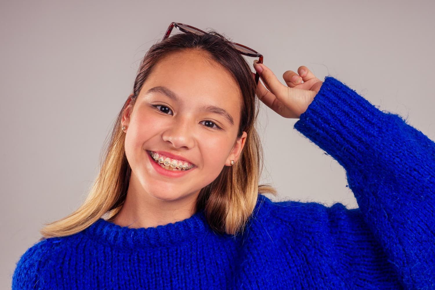 A girl with braces, smiling in a blue sweater, holds eyeglasses above her head for Stellar Orthodontics in San Marcos, CA.