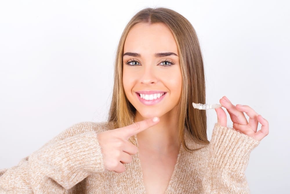 A smiling woman points to her teeth, holding a clear aligner with one hand for Stellar Orthodontics in San Marcos, CA - Can Invisalign Fix Overbite in San Marcos, CA.