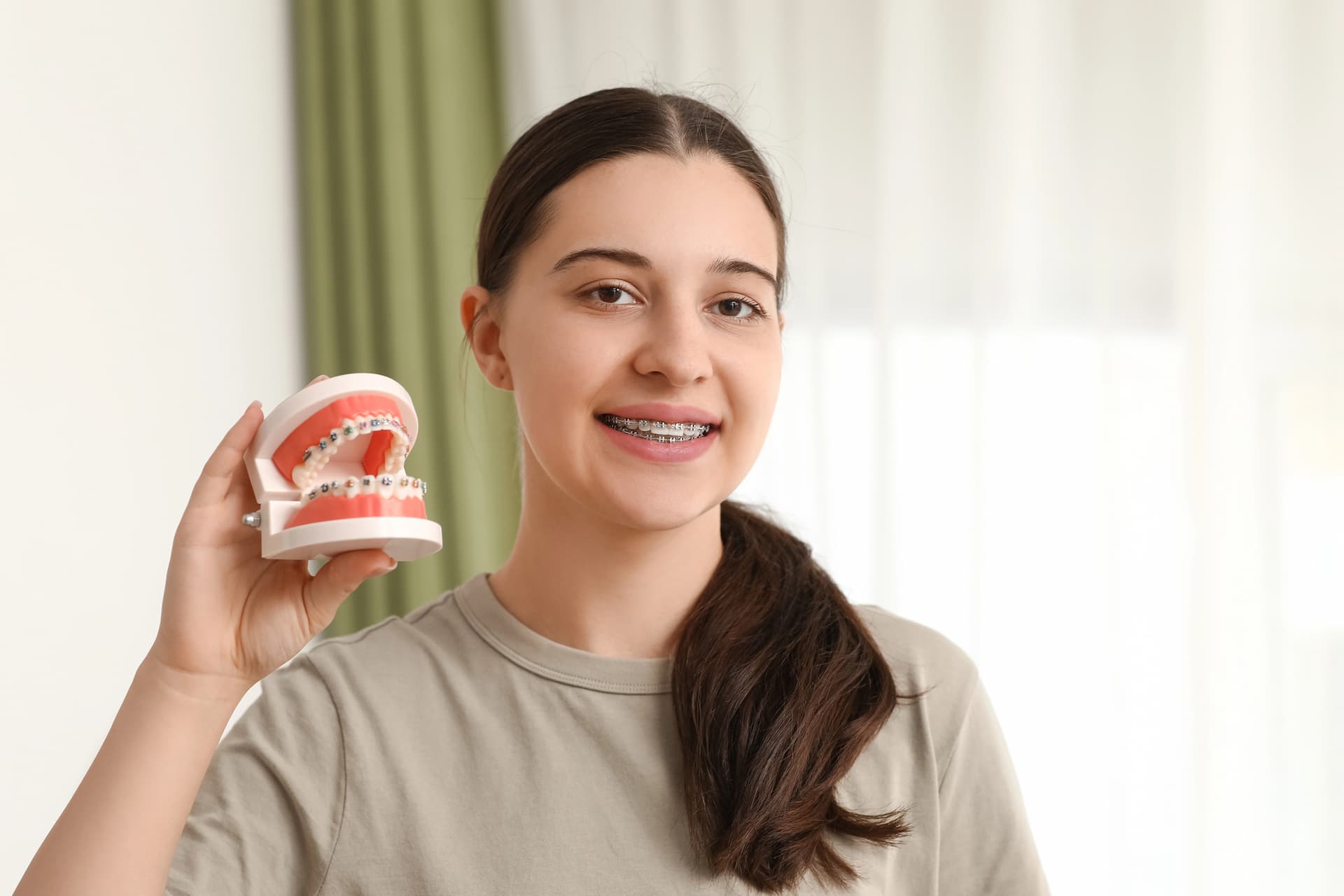 Smiling young woman with metal braces holds a dental model, representing Stellar Orthodontics in San Marcos, CA, against a bright background.