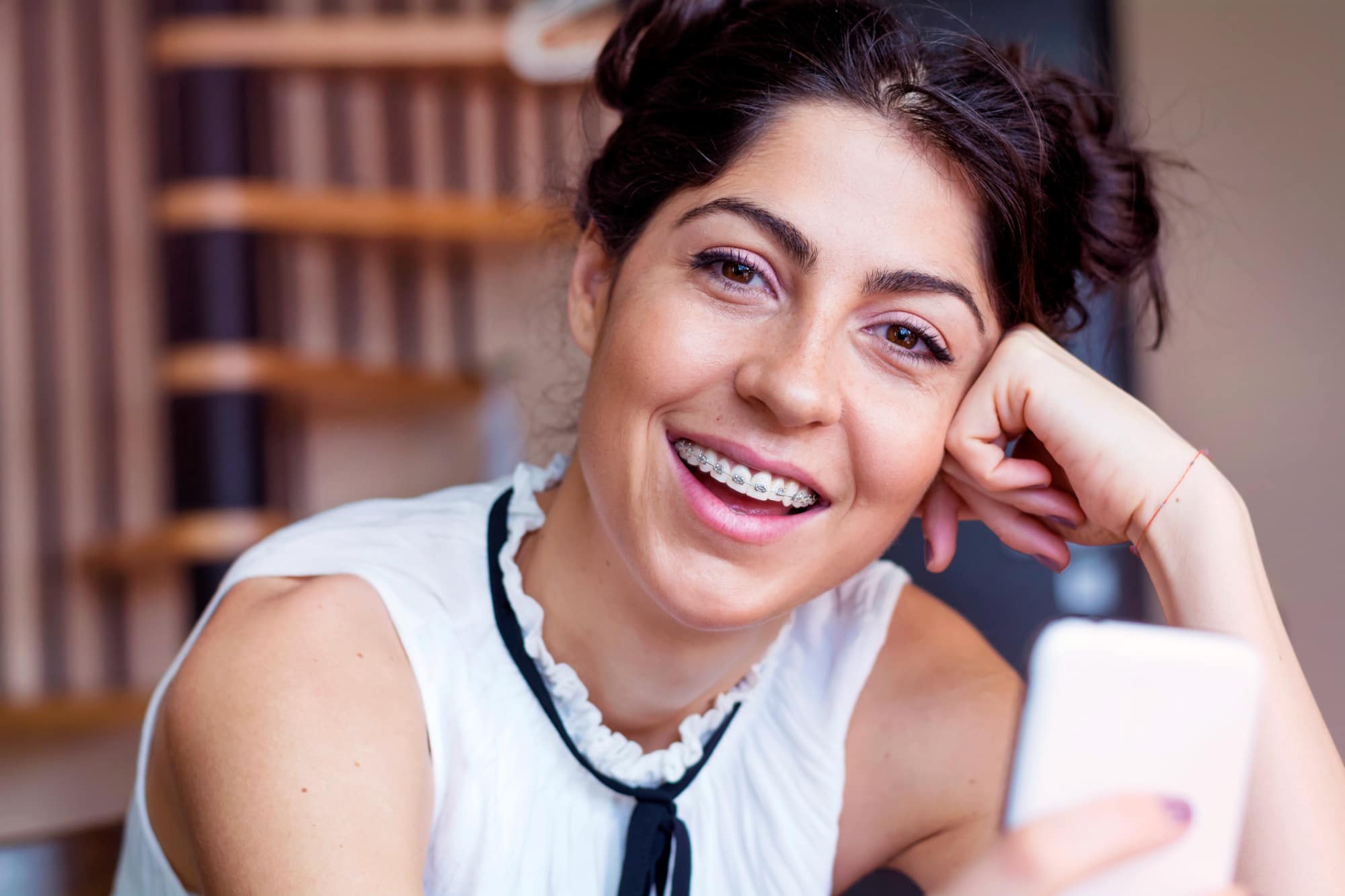 Smiling young woman with metal braces holds a smartphone indoors, representing Stellar Orthodontics in San Marcos, CA.
