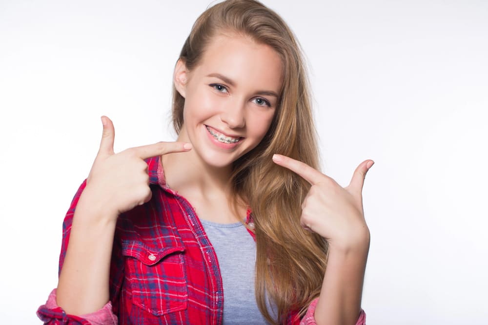 Stellar Orthodontics A smiling teen with long blonde hair in a red plaid shirt points at her braces, highlighting affordable braces cost at Stellar Orthodontics in San Marcos, CA.