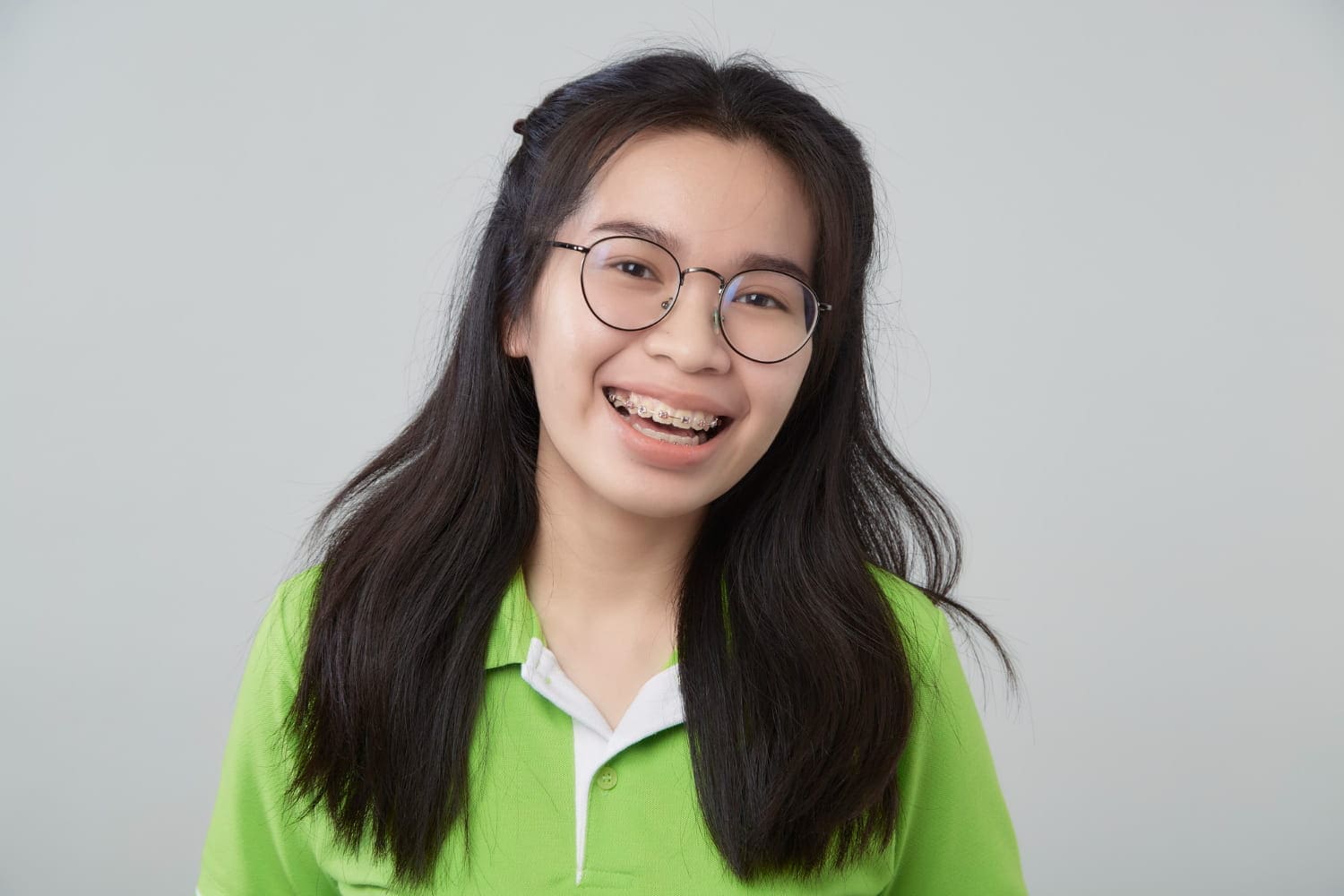 Smiling teen with long dark hair, glasses, and metal braces at Stellar Orthodontics in San Marcos, CA; green shirt, gray backdrop.